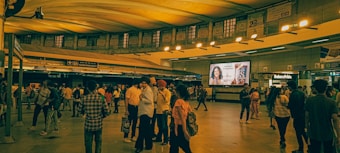 A crowded metro station with people standing, walking, and waiting. The ceiling features an arched design with bright yellow lighting. Advertisements are displayed on digital screens, and various signs provide directions and information.