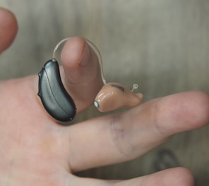 A hand is holding a hearing aid, which consists of a small beige earpiece connected by a thin wire to a black device.