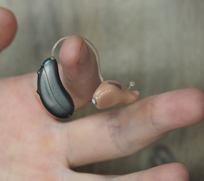A hand is holding a hearing aid, which consists of a small beige earpiece connected by a thin wire to a black device.