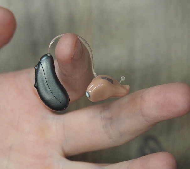 A hand is holding a hearing aid, which consists of a small beige earpiece connected by a thin wire to a black device.