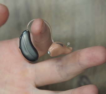 A hand is holding a hearing aid, which consists of a small beige earpiece connected by a thin wire to a black device.