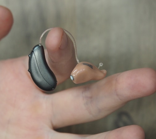 A hand is holding a hearing aid, which consists of a small beige earpiece connected by a thin wire to a black device.