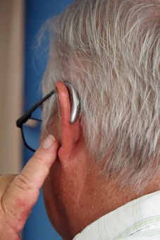 An elderly person with grey hair uses a hearing aid. The person is wearing glasses and has their finger touching the hearing aid behind their ear.