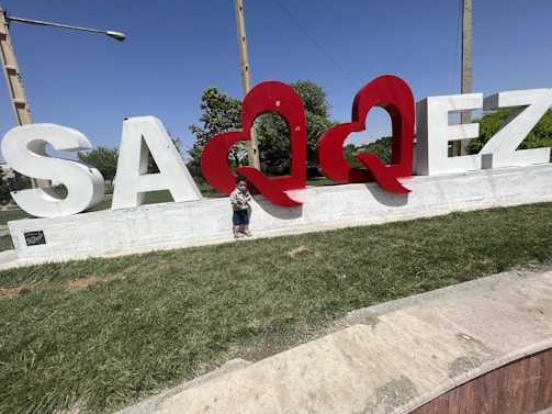 Large white letters with two prominent red heart shapes spell out 'SAQQEZ' displayed on a raised concrete platform bordered by grass. A young child is standing near the red heart shapes. In the background, there are green trees and a clear blue sky.