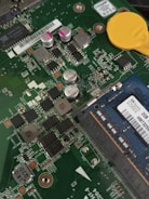 A close-up view of a computer motherboard with various components including capacitors, resistors, and a RAM module. The circuitry is intricate, and various labels and numbers are visible on the chips. A yellow battery is positioned at the top, and there's a blue RAM card labeled with information such as 2GB and Korea.