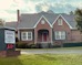 A traditional red brick building with a gabled roof and white-framed windows. The front entrance features an arched doorway and is flanked by well-maintained shrubs. A sign in the foreground reads 'Morris & Dean, Trial Lawyers & Counselors,' displaying the phone number and a digital clock showing 6:31.
