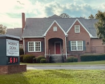 A traditional red brick building with a gabled roof and white-framed windows. The front entrance features an arched doorway and is flanked by well-maintained shrubs. A sign in the foreground reads 'Morris & Dean, Trial Lawyers & Counselors,' displaying the phone number and a digital clock showing 6:31.