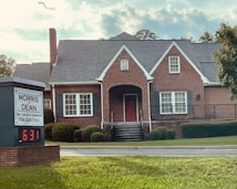 A traditional red brick building with a gabled roof and white-framed windows. The front entrance features an arched doorway and is flanked by well-maintained shrubs. A sign in the foreground reads 'Morris & Dean, Trial Lawyers & Counselors,' displaying the phone number and a digital clock showing 6:31.