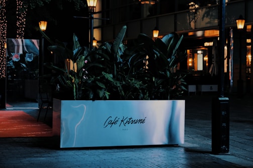 A stylish and modern planter with lush green foliage is placed outside a cafe or restaurant, which is softly illuminated by warm street lamps. The planter is marked 'Café Kitsuné Paris' and there are reflective surfaces with a cozy, urban ambiance.