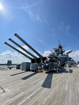 A military ship with large cannons is docked on a port with a wooden deck. A helicopter is positioned on the deck, surrounded by ropes and barriers. The background features a clear blue sky with some clouds and a distant view of a tall building near the water.