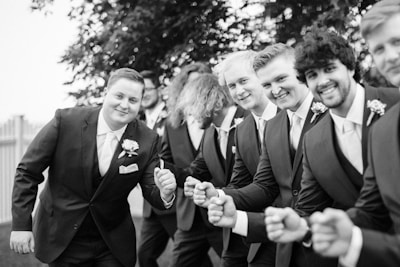 A group of men dressed in formal suits with boutonnieres are posing together outdoors. They appear to be happy, smiling, and making enthusiastic gestures with their fists as if celebrating or cheering. The background includes some greenery, such as trees, and a glimpse of a fence on the left side.