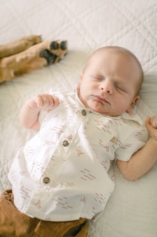 A sleeping baby wearing a patterned white shirt lies peacefully on a quilted bedspread. A dog's paw is visible in the background, adding a gentle presence to the scene.