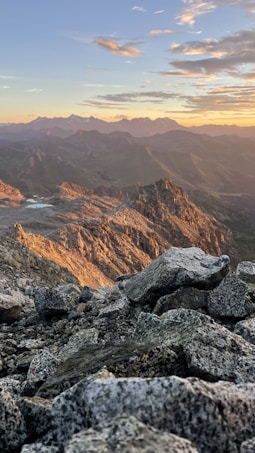 A rugged mountain landscape at sunset, with rocky foreground leading to distant peaks silhouetted against a colorful sky. The sunlight casts a warm glow over the terrain, highlighting the textures and contours of the rocks.