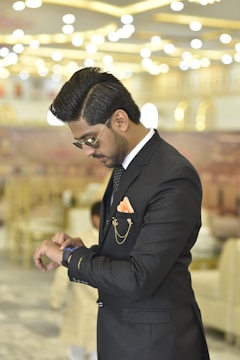 A man in a formal black suit is looking at his wristwatch in an indoor setting with modern, elegant lighting. He wears glasses and his hair is neatly styled. The background is softly blurred, showcasing a sophisticated and opulent interior design.