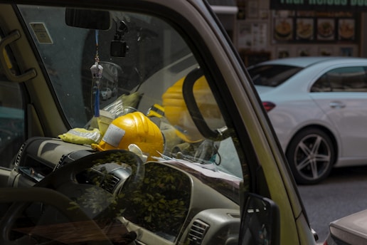 A close-up view of the interior of a vehicle, focusing on the dashboard area where two yellow construction helmets are resting. The sunlight entering through the windows creates reflections on the windscreen, and there is a hanging ornament with a blue tassel from the rearview mirror. Outside the vehicle, a white car is partially visible, driving along the street.