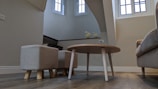 A cozy indoor space with modern furniture, including a round wooden table and small upholstered stools on a wooden floor. The room has light beige walls and features bright natural light coming through tall windows. Decorative items like two bird figurines are placed on a shelf near the stairs.