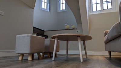 A cozy indoor space with modern furniture, including a round wooden table and small upholstered stools on a wooden floor. The room has light beige walls and features bright natural light coming through tall windows. Decorative items like two bird figurines are placed on a shelf near the stairs.
