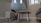 A cozy indoor space with modern furniture, including a round wooden table and small upholstered stools on a wooden floor. The room has light beige walls and features bright natural light coming through tall windows. Decorative items like two bird figurines are placed on a shelf near the stairs.