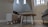 A cozy indoor space with modern furniture, including a round wooden table and small upholstered stools on a wooden floor. The room has light beige walls and features bright natural light coming through tall windows. Decorative items like two bird figurines are placed on a shelf near the stairs.