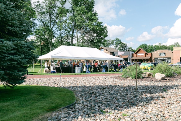 A large white tent is set up outdoors on a grassy area surrounded by trees and commercial buildings. Inside the tent, people are gathered for what looks like a social or community event. The foreground features a rock garden with greenery, and the sky is clear with scattered clouds.