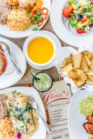 A variety of Mexican dishes are arranged on a table, including plates of rice, beans, enchiladas, and a fresh salad alongside a bowl of cheese dip and a basket of tortilla chips. A green beverage is served in a glass with ice. A menu from Alicia's Mexican Grille is visible on the table.