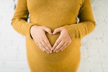 A person wearing a mustard-colored knitted sweater is making a heart shape with their hands over their belly.