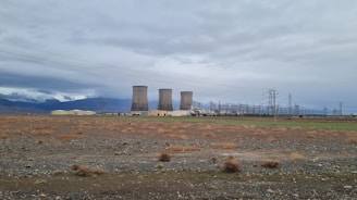 Several large cooling towers are situated in a wide, open field under a cloudy sky. The ground is mostly barren with sparse vegetation and some power lines are visible. Mountains can be seen in the distance, partially covered by clouds.