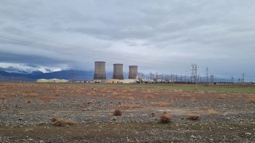 Several large cooling towers are situated in a wide, open field under a cloudy sky. The ground is mostly barren with sparse vegetation and some power lines are visible. Mountains can be seen in the distance, partially covered by clouds.