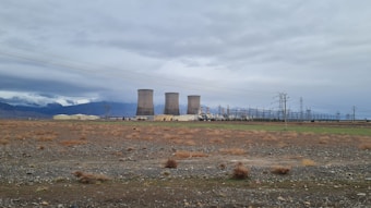 Several large cooling towers are situated in a wide, open field under a cloudy sky. The ground is mostly barren with sparse vegetation and some power lines are visible. Mountains can be seen in the distance, partially covered by clouds.