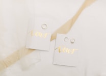 A pair of small booklets labeled 'His Vows' and 'Her Vows' placed on a white surface. Each booklet has a gold-colored title, and next to each booklet, there is a wedding ring. A sheer piece of fabric is draped decoratively over part of the scene.