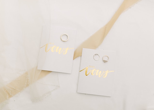 A pair of small booklets labeled 'His Vows' and 'Her Vows' placed on a white surface. Each booklet has a gold-colored title, and next to each booklet, there is a wedding ring. A sheer piece of fabric is draped decoratively over part of the scene.