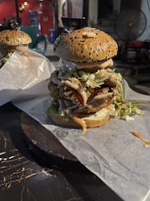 A large, stacked burger on a round plate lined with parchment paper. The burger features a sesame seed bun and is filled with layers of crispy fried chicken, shredded lettuce, and drizzles of sauce. The setting is likely a casual dining environment with another similar burger partially visible in the background.