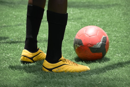 A pair of legs wearing black socks and yellow soccer shoes stands near a red and black soccer ball on a grass field.
