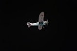 A vintage-style airplane with a blue and white color scheme is flying against a dark, almost black background. The registration on the aircraft's tail is visible. The plane has a high wing design and two propellers near the nose.