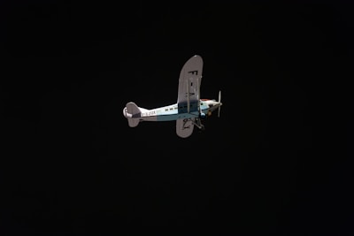 A vintage-style airplane with a blue and white color scheme is flying against a dark, almost black background. The registration on the aircraft's tail is visible. The plane has a high wing design and two propellers near the nose.
