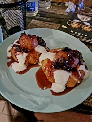 Two dessert pastries topped with a red sauce, dark berries, and white cream are served on a light blue plate. The setting includes a woven surface, a drink menu, a small bottle of water, a metal basket, and a glass in the background.