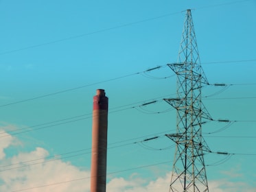 A large smokestack is standing next to a tall electrical transmission tower under a clear blue sky. The smokestack is cylindrical and has a reddish-brown color. The tower is made of metal and has multiple cables extending from it.