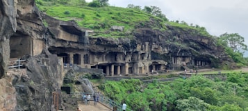 A series of ancient rock-cut caves are carved into a hillside, surrounded by lush greenery. The caves exhibit intricate architectural details with pillars and entrances. Several people are walking along the path in front of the caves, which are set within a rocky landscape.