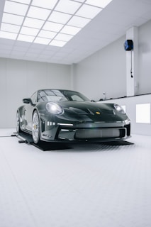 A sleek, luxury sports car with a glossy black finish is positioned in a modern, well-lit showroom. The car features distinctive curves and aerodynamic design, emphasizing its high-performance capabilities. The environment is minimalistic, with white walls and ceiling lights creating a clean and futuristic ambiance.
