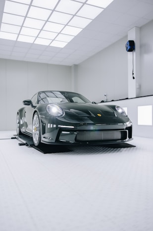 A sleek, luxury sports car with a glossy black finish is positioned in a modern, well-lit showroom. The car features distinctive curves and aerodynamic design, emphasizing its high-performance capabilities. The environment is minimalistic, with white walls and ceiling lights creating a clean and futuristic ambiance.