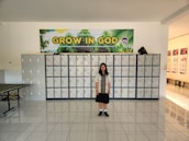 A girl stands in front of a wall of lockers. Above the lockers is a large banner with the words 'GROW IN GOD' and an image of greenery. The floor is tiled and there is a table to the left.