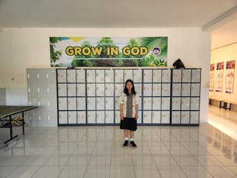 A girl stands in front of a wall of lockers. Above the lockers is a large banner with the words 'GROW IN GOD' and an image of greenery. The floor is tiled and there is a table to the left.