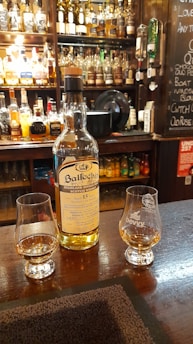 A bottle of Ballechin Highland Single Malt Scotch Whisky, aged 15 years, is placed on a wooden bar counter. Two glasses containing whisky are positioned next to the bottle. In the background, there is a wide variety of liquor bottles on shelves behind the bar, alongside a chalkboard with menu items. The setting is warmly lit, creating a cozy atmosphere.