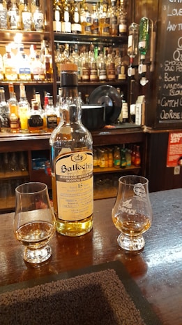 A bottle of Ballechin Highland Single Malt Scotch Whisky, aged 15 years, is placed on a wooden bar counter. Two glasses containing whisky are positioned next to the bottle. In the background, there is a wide variety of liquor bottles on shelves behind the bar, alongside a chalkboard with menu items. The setting is warmly lit, creating a cozy atmosphere.