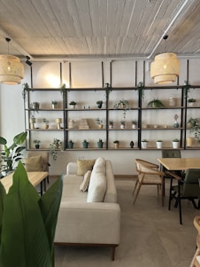 A cozy living room featuring minimalist beige furniture, soft glowing lamps, textured gray walls, and lush green indoor plants.