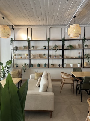 A cozy, minimalist interior featuring a beige sofa with several cushions, surrounded by wooden tables and chairs. A large shelf unit with various potted plants, decorative items, and small lamps lines the back wall, adding a touch of greenery and warmth. The ceiling is made of wooden panels with two wicker pendant lights hanging down, emitting a soft glow.