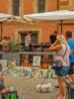 A street scene with people observing a display of various paintings set on the pavement against the backdrop of a historic building with large windows. The paintings include animal portraits and human figures. A couple stands in the foreground with a small white dog on a leash.