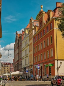 A European street scene featuring colorful buildings with intricate architectural details, including red, yellow, and beige facades. People are walking along the cobblestone street, and outdoor seating is visible under white umbrellas. The sky is clear and blue, adding to the vibrant atmosphere.