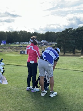 Two people on a golf course, one wearing a pink top and black pants, the other a white and blue outfit with golf equipment. A large crowd and trees are visible in the background under a partly cloudy sky.