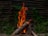 A stack of wooden logs burning with bright orange flames that rise into the air. The fire is situated in an outdoor setting, surrounded by lush green vegetation. In the background, there is a rustic wooden fence made from horizontal wooden slats, adding to the natural environment.